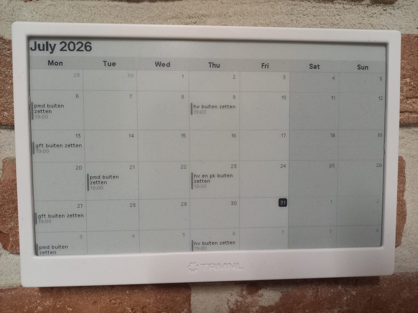 Een e-inkscherm aan een bakstenen muur, met de maandkalender van juli 2026. In de vakjes staat pmd buiten zetten, gft buiten zetten en hv en pk buiten zetten, telkens met 19:00 erbij.